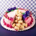 A round pink frosted cake on a white scalloped paper plate, decorated with purple frosting rosettes, bright pink piped borders, multicolored sprinkles, and a cluster of pale yellow rolled pastry pieces on top. The cake is photographed against a blue-and-white checkered backdrop on a dark purple surface.