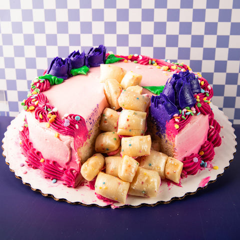 A round pink frosted cake on a white scalloped paper plate, decorated with purple frosting rosettes, bright pink piped borders, multicolored sprinkles, and a cluster of pale yellow rolled pastry pieces on top. The cake is photographed against a blue-and-white checkered backdrop on a dark purple surface.