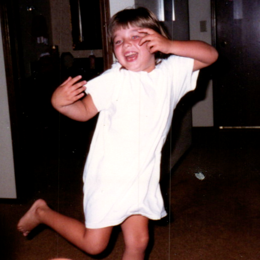 A young child wearing a white short-sleeve t-shirt dress, captured mid-jump in a dimly lit indoor room. The child is smiling and covering one eye with their hand.