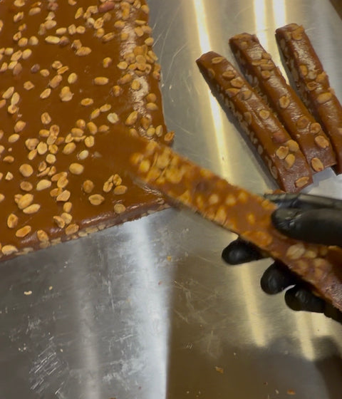 A close-up of a hand wearing a black glove slicing a long, glossy caramel candy bar topped with chopped peanuts on a stainless steel surface, with more candy bars and a large sheet of caramel candy visible in the background.