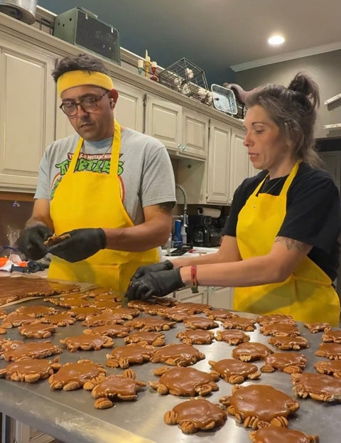 Two people in yellow aprons and black gloves work at a stainless steel table, shaping and making tutles in a kitchen setting.