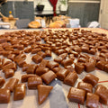 A close-up, angled view of many brown, glossy, pieces of salted carmel arranged on a baking sheet or tray, with a blurred indoor room in the background featuring a red curtain and a sign that says DINER.