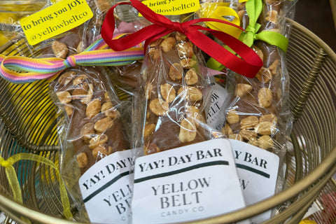 Several individually wrapped Yellow Belt candy bars tied with colorful ribbons and placed in a gold wire basket. The clear wrapping shows the caramel-coated yellowpeanut candy inside, with white labels reading 'YAY! DAY BAR' and 'YELLOW BELT'.