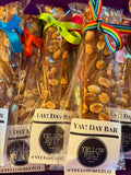 Several packaged yellow candy bars with nuts, tied with colorful ribbons, displayed in a row. The labels read 'YAY! DAY BAR' and 'YELLOW BELT JOY CO.